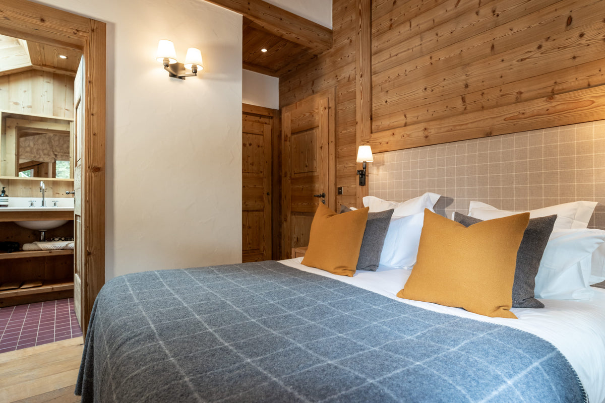 A cozy bedroom interior with a large bed dressed in a blue-gray plaid blanket, white bedding, mustard yellow and gray accent pillows, and a wood-paneled wall. A doorway on the left reveals a bathroom with a sink and mirror.