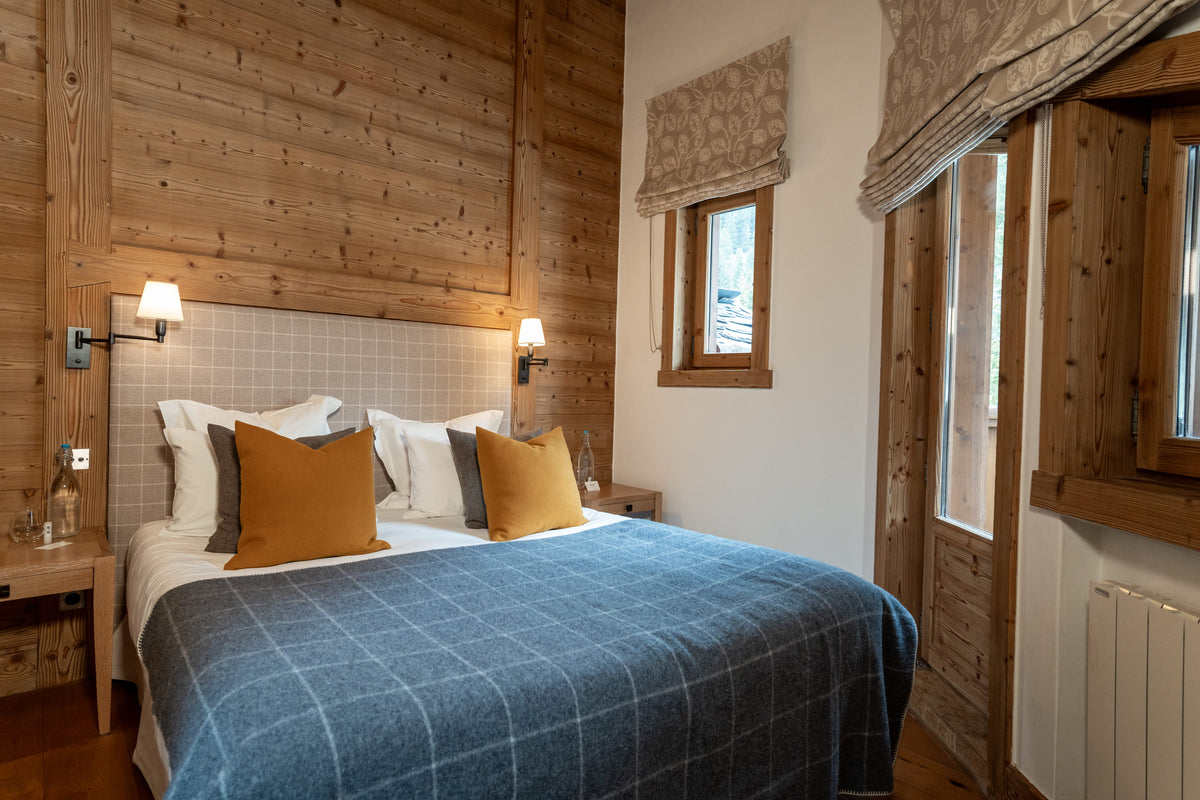 A cozy bedroom with a neatly made bed featuring a blue plaid blanket, mustard yellow and gray accent pillows, set against a rustic wood-paneled wall with bedside lamps and a small window.