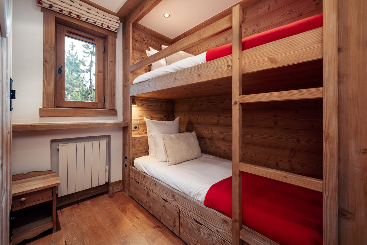 Alpine-style wooden bunk bed with two single beds, dressed in white linens and bright red accent sheets, set against a wood-paneled wall in a cozy room with a window and small wooden side table.