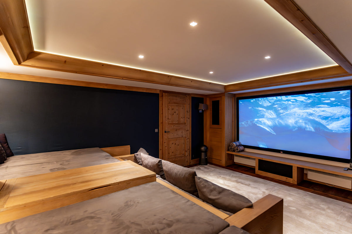 A modern home theater room with a large projection screen displaying a snowy mountain scene, built into a wooden wall with a dark blue accent panel. In the foreground, a low wooden sofa with gray cushions faces the screen, and the room is illuminated by warm recessed lighting and LED strip lights in the ceiling.