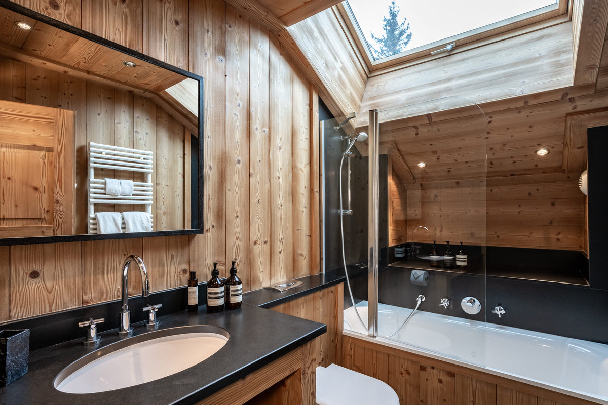 Luxury wood-paneled bathroom with a black countertop, white sink, large mirror, glass shower enclosure, and white bathtub under a skylight.