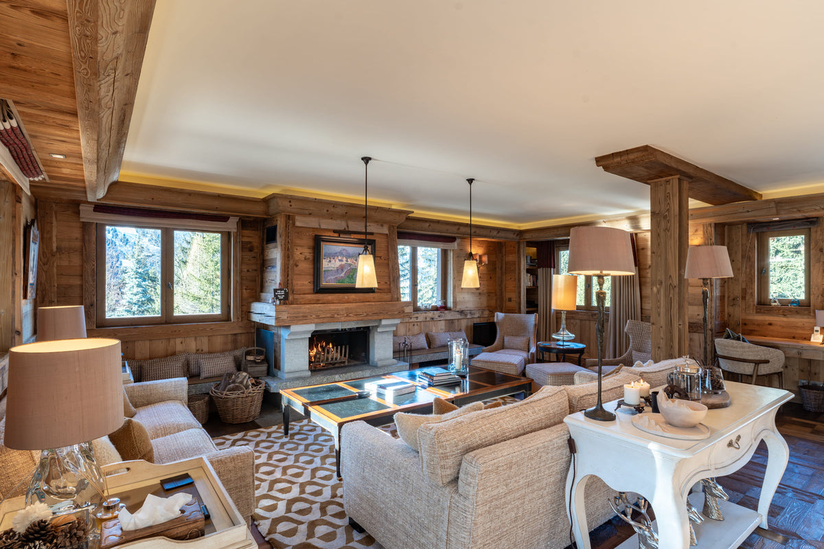 A luxurious ski chalet living room featuring rustic wood walls and ceiling beams, a central fireplace, a patterned area rug, beige upholstered sofas, and multiple table lamps.