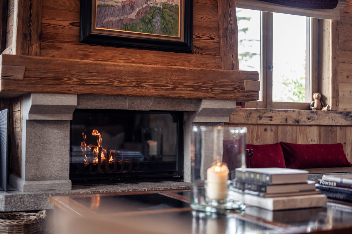 A cozy interior scene featuring a stone and wood fireplace with a visible fire, set within a rustic wooden wall. In the foreground, a glass candle holder and stacked books sit on a table, while a window in the background shows red cushions and a small figurine.
