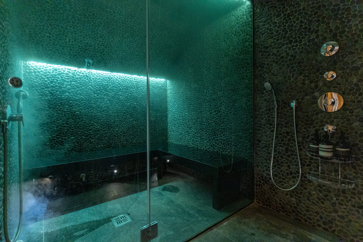 A modern glass shower enclosure with a dark stone-tiled interior and green-tinted ambient lighting, showing a built-in bench, handheld shower, and decorative wall mirrors.