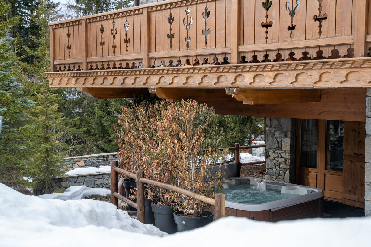 A wooden chalet-style outdoor hot tub deck in a snowy mountain setting, featuring a carved wooden balcony with decorative metal railings and a stone-and-wood exterior.