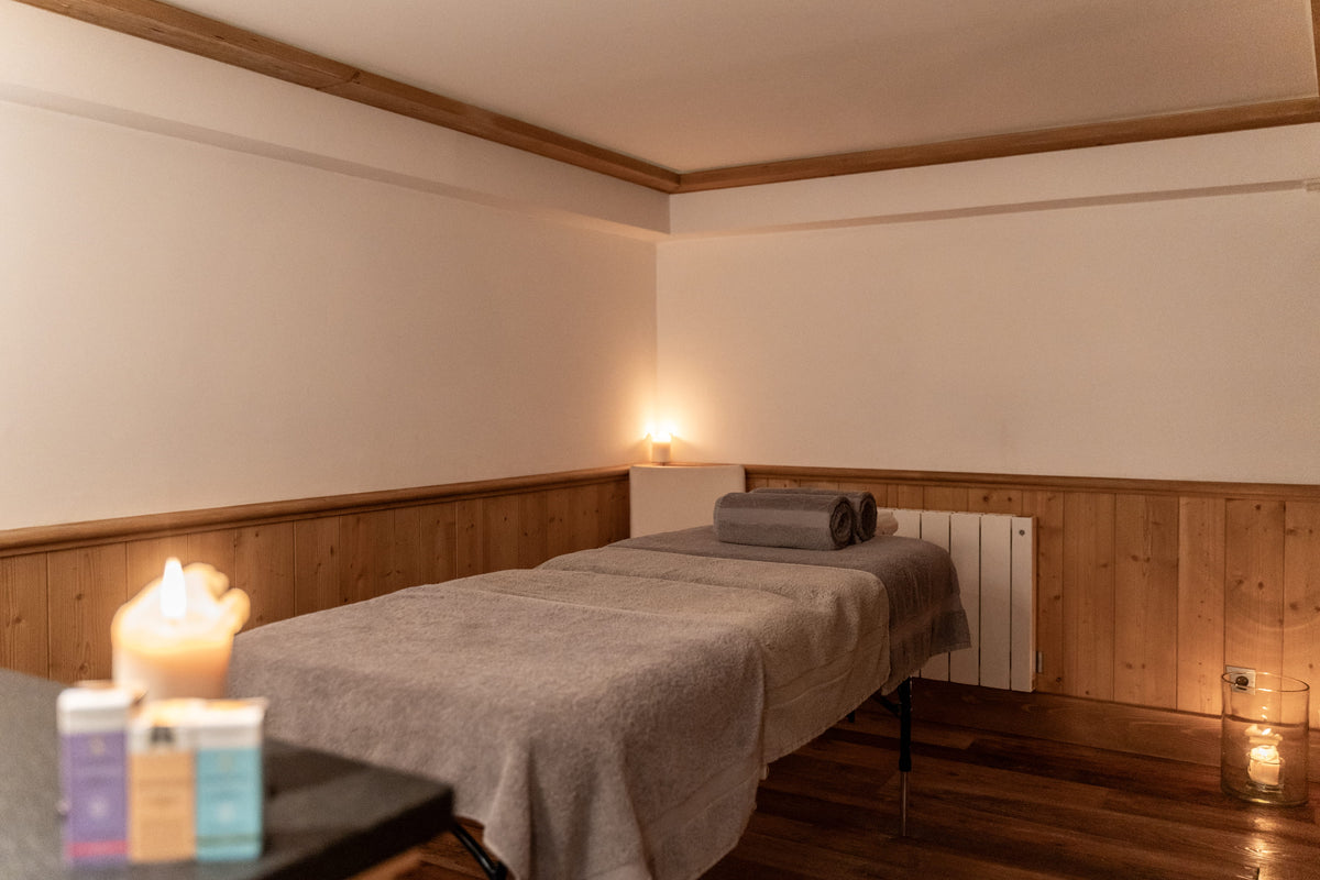 A spa treatment bed with a gray blanket and rolled towel in a warmly lit room with wood paneling, candles, and small product boxes in the foreground.