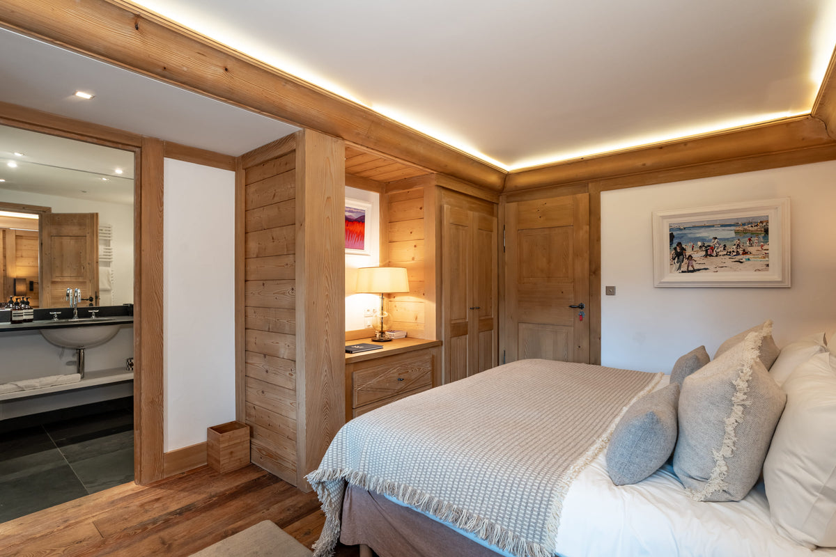 A modern ski chalet bedroom featuring a large bed with white and gray bedding, light wood walls, and a built-in wooden wardrobe. An open doorway on the left reveals a bathroom with a white sink and dark tiled floor.