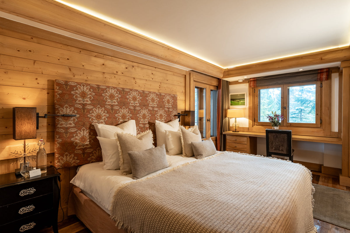 A cozy bedroom with a large bed dressed in cream bedding and multiple pillows, set against a wood-paneled wall with a patterned headboard. A matching desk and chair sit by a window with daylight coming in.