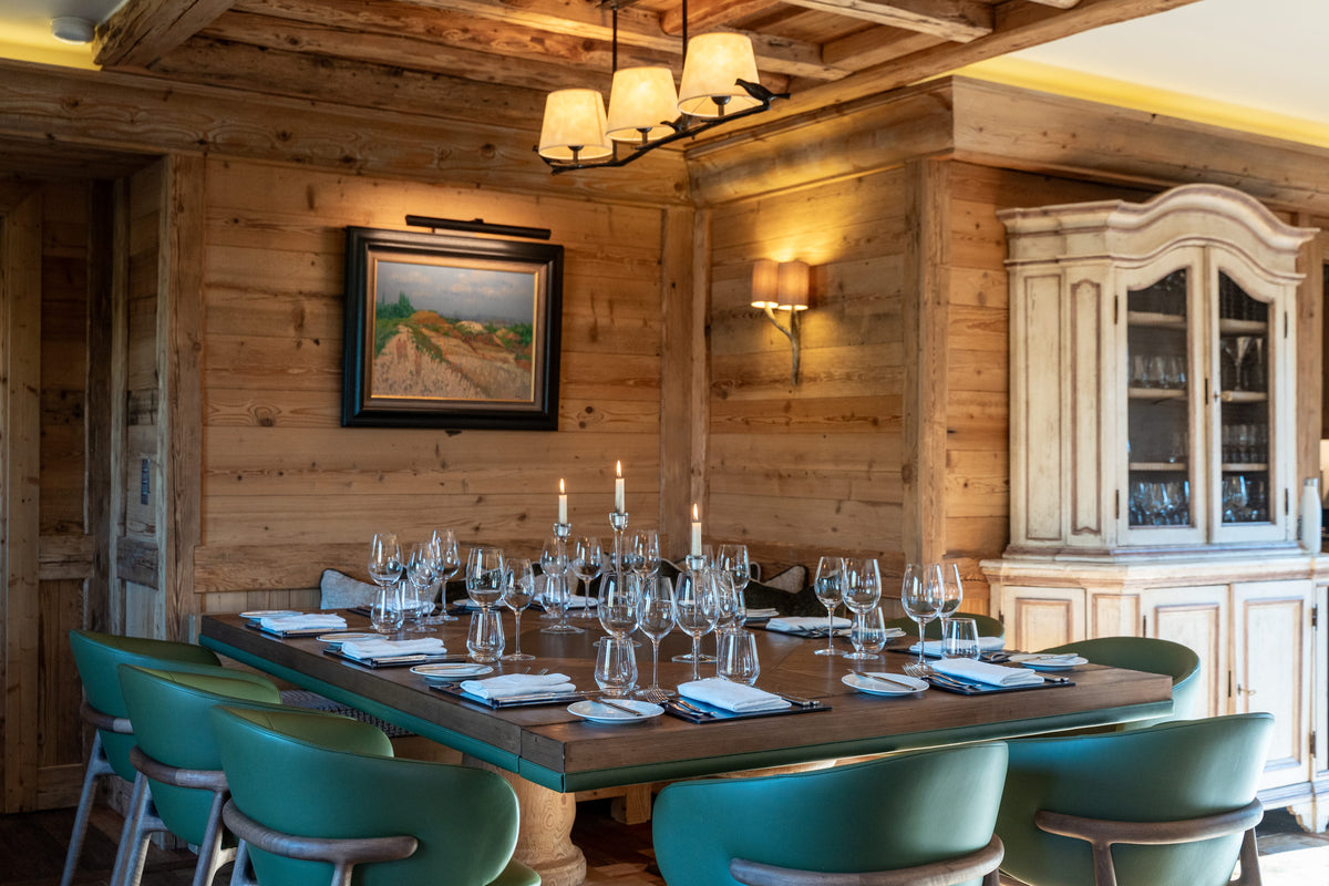 A rustic dining room with wood-paneled walls, a long wooden table set with wine glasses and candles, teal chairs, and a carved wooden hutch.
