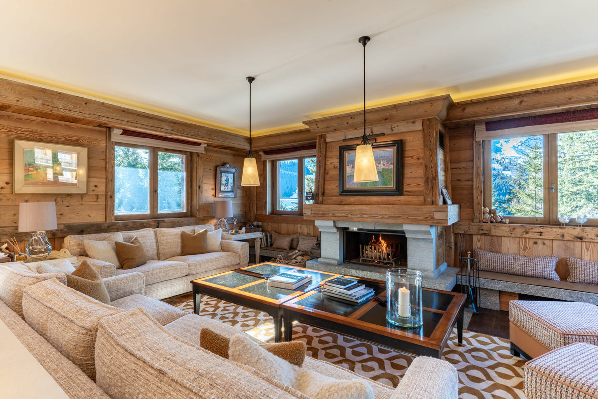 A cozy, rustic mountain-style living room with wood-paneled walls, a stone fireplace, and multiple beige upholstered sofas arranged around a patterned rug and dark wood coffee table.