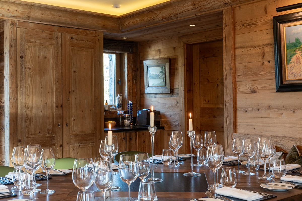 A rustic wooden dining room with a long table set with wine glasses, candles, and white tablecloths, surrounded by warm wood-paneled walls and cabinetry.