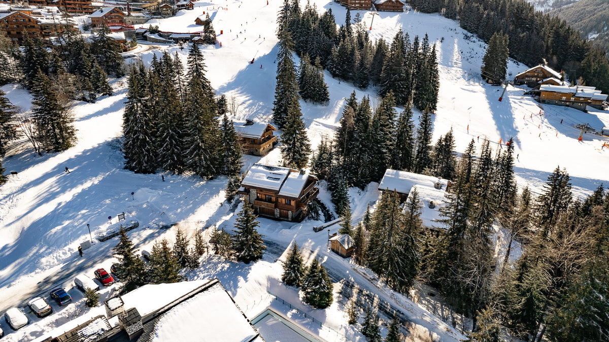 Aerial view of a snowy mountain village with chalets, evergreen trees, ski slopes, and parked cars in a winter alpine setting.