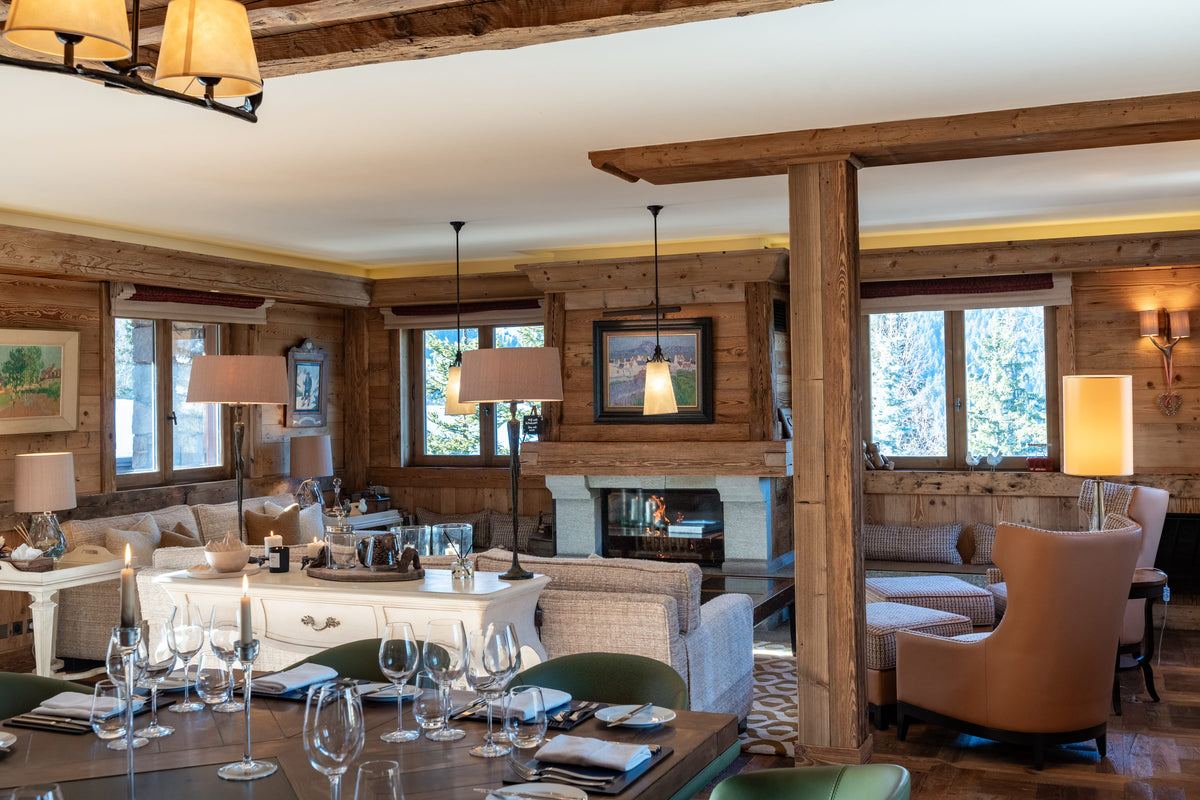 A rustic lodge-style living room with exposed wooden beams, a fireplace, and multiple upholstered seating areas. A dining table with wine glasses is visible in the foreground, and the room is warmly lit by hanging and table lamps.