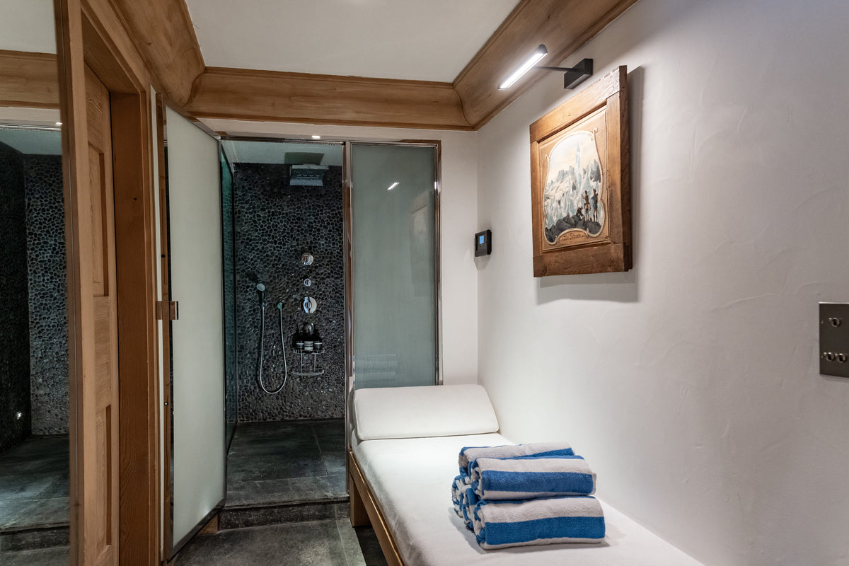 A minimalist spa-style room with a wooden-framed massage table, white walls, a framed artwork, and a walk-in shower area in the background. A stack of blue and white towels sits on the table.