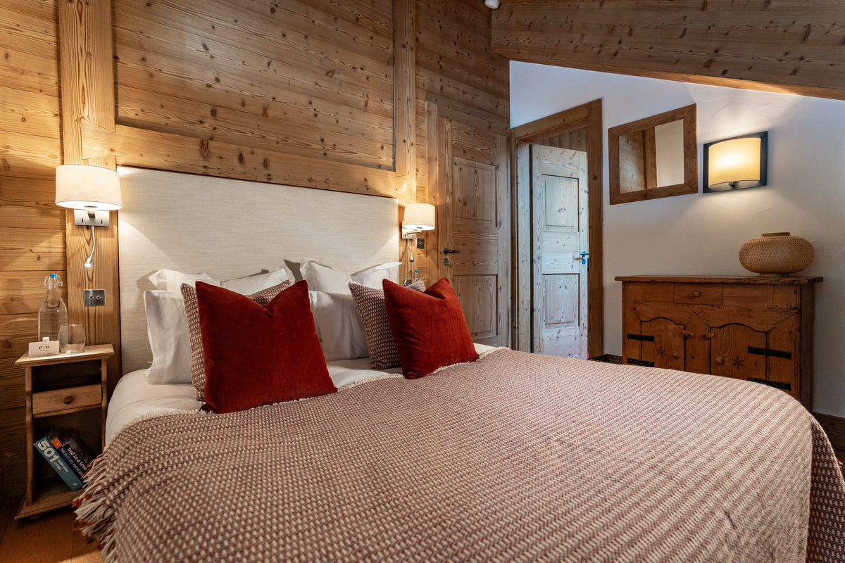 A cozy bedroom with a large bed dressed in a textured beige blanket and red accent pillows, set against rustic wood-paneled walls with bedside lamps and a wooden dresser.