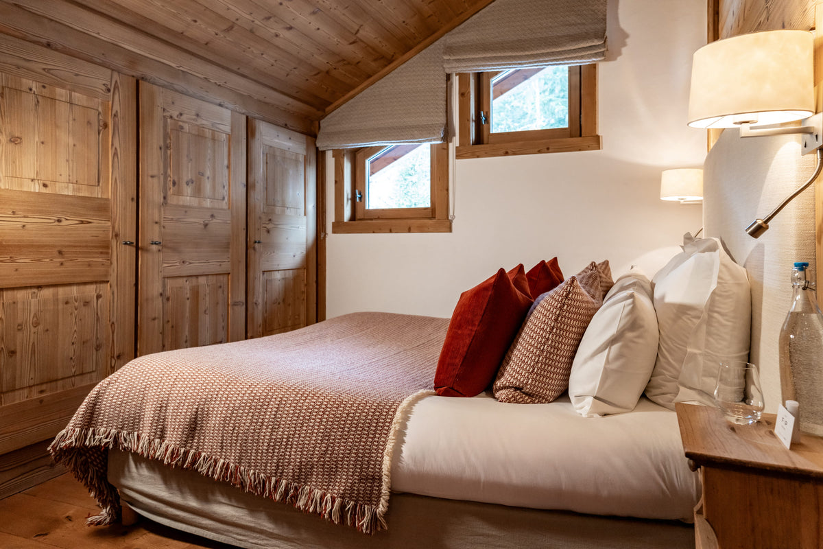 A cozy bedroom interior with a neatly made bed dressed in cream bedding, a rust-brown fringed throw blanket, and several pillows including a prominent red one. The room features light wood paneled walls, a matching wooden wardrobe, a slanted ceiling, and a bedside table with a lit wall lamp.