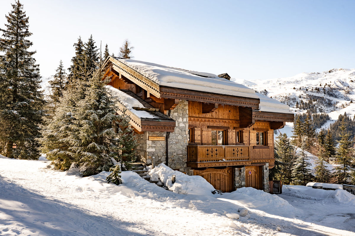 A traditional alpine chalet with snow-covered roofs and wooden balconies, surrounded by snow-laden pine trees in a mountain landscape under a clear blue sky.