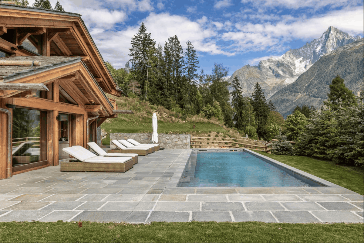 A luxury mountain chalet with a stone patio, outdoor swimming pool, two white lounge chairs, and a stunning backdrop of pine trees and snow-capped peaks under a blue sky.