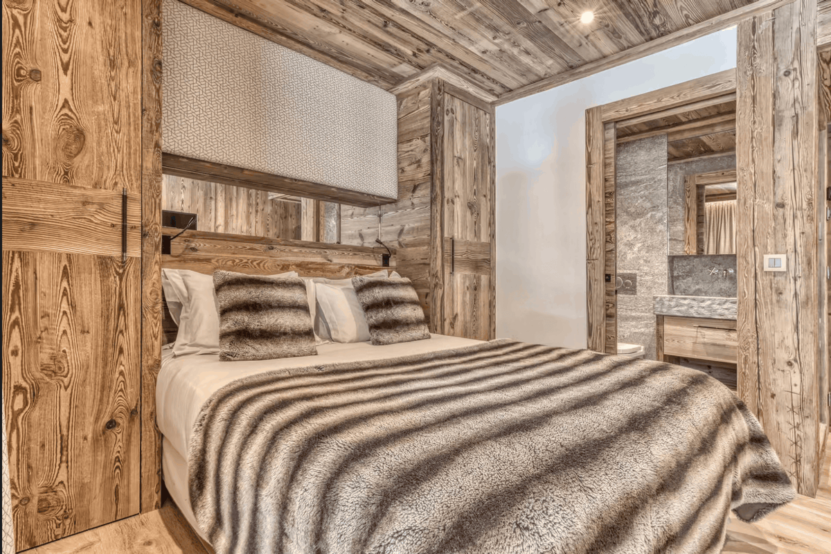 A rustic bedroom interior with a large bed dressed in white bedding and a brown-and-white striped animal-print throw blanket, surrounded by wood-paneled walls and ceiling. A doorway to a stone bathroom is visible on the right.
