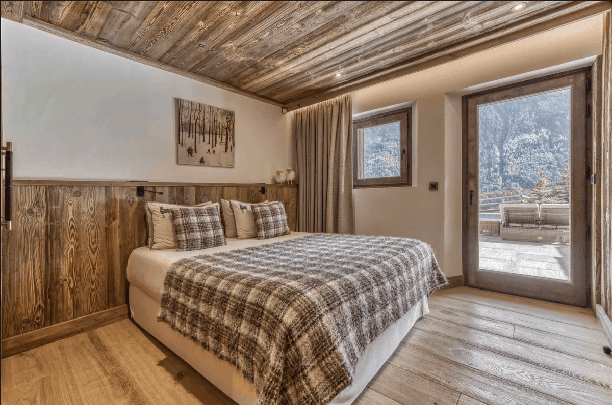 A rustic bedroom with a large bed dressed in beige and brown plaid bedding, set against a wood-paneled wall. The room features a wooden ceiling, light wood floors, and a glass door opening to a snowy mountain landscape.