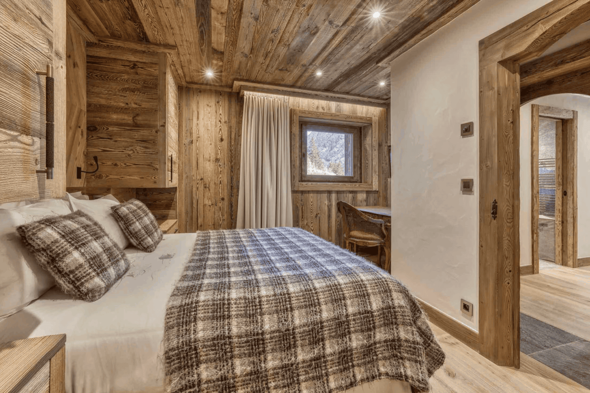 A cozy bedroom with rustic wooden walls and ceiling, featuring a bed dressed in white linens and a brown-and-gray plaid blanket, matching pillows, a small desk by a window, and warm ambient lighting.