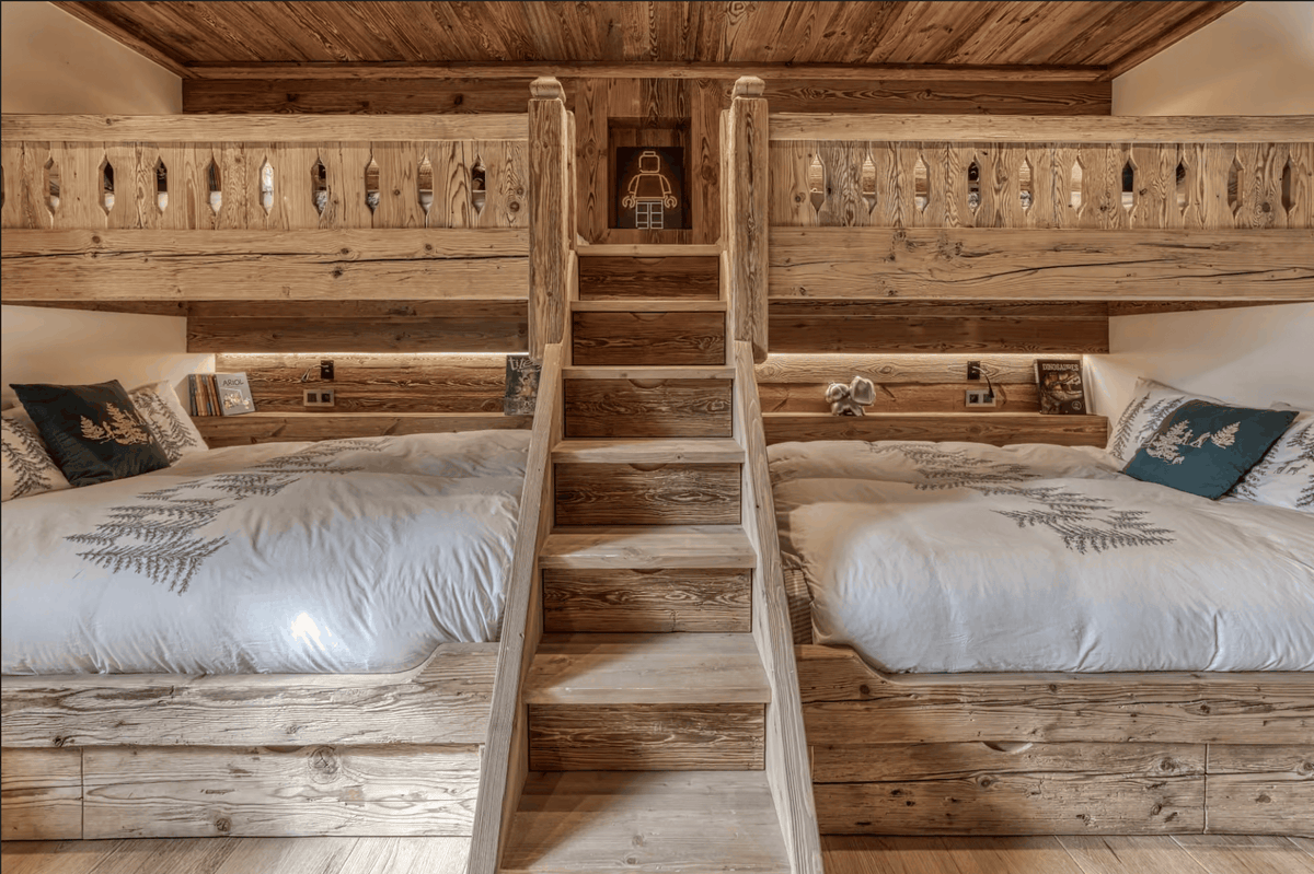 A rustic wooden loft bed with two single beds on either side of a central wooden ladder, featuring white bedding with gray fern patterns and dark blue accent pillows.