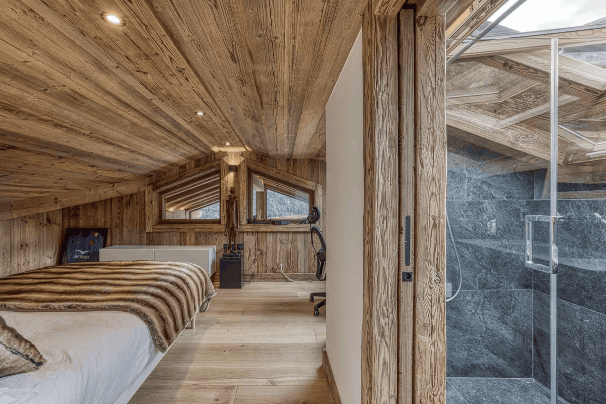 A rustic mountain chalet bedroom featuring a bed with a brown and beige animal-print throw blanket, a glass shower enclosure with a stone-tiled shower room, and a sloped wooden ceiling.