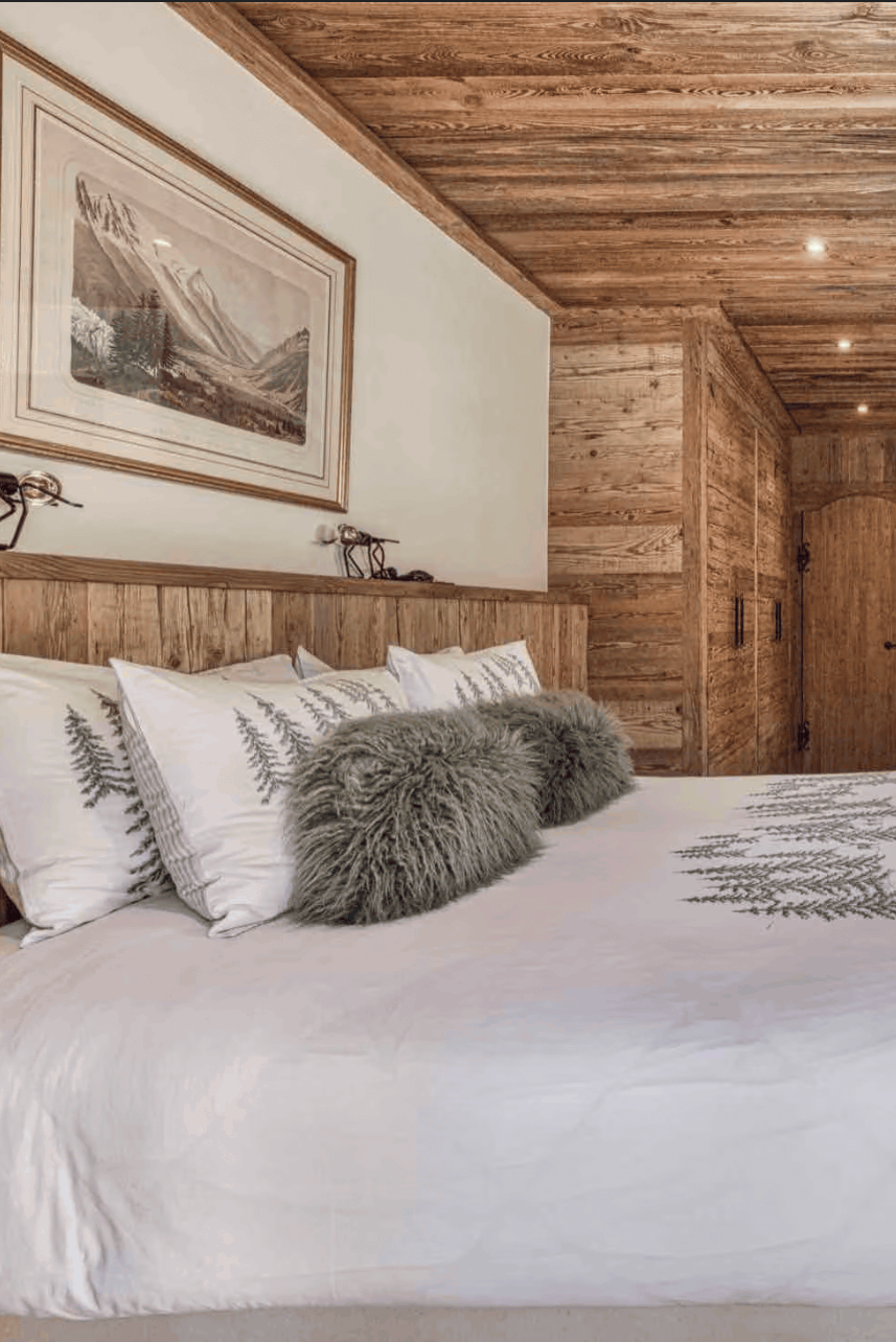 A rustic mountain chalet bedroom featuring a large bed with white bedding and gray accent pillows, set against warm wood-paneled walls and ceiling with a window and chair nearby.