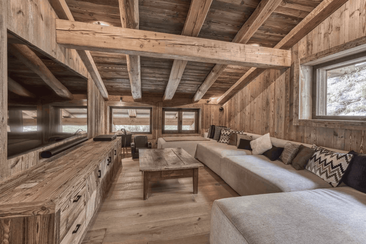 A rustic mountain chalet living room featuring a large beige sectional sofa with patterned pillows, a wooden coffee table, a built-in TV on a wood cabinet, and a sloped ceiling with exposed wooden beams.