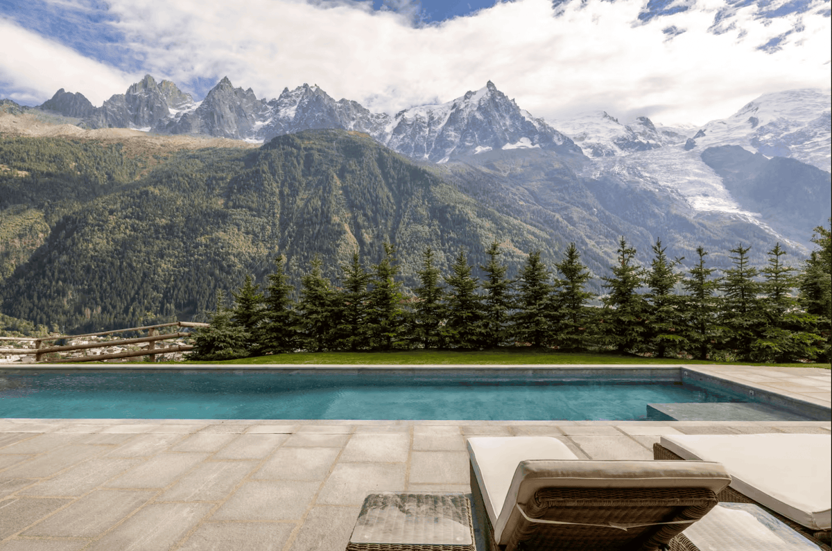 A luxury outdoor pool with a stone deck and lounge chair, set against a backdrop of dense green pine trees and majestic snow-capped mountain peaks under a cloudy sky.