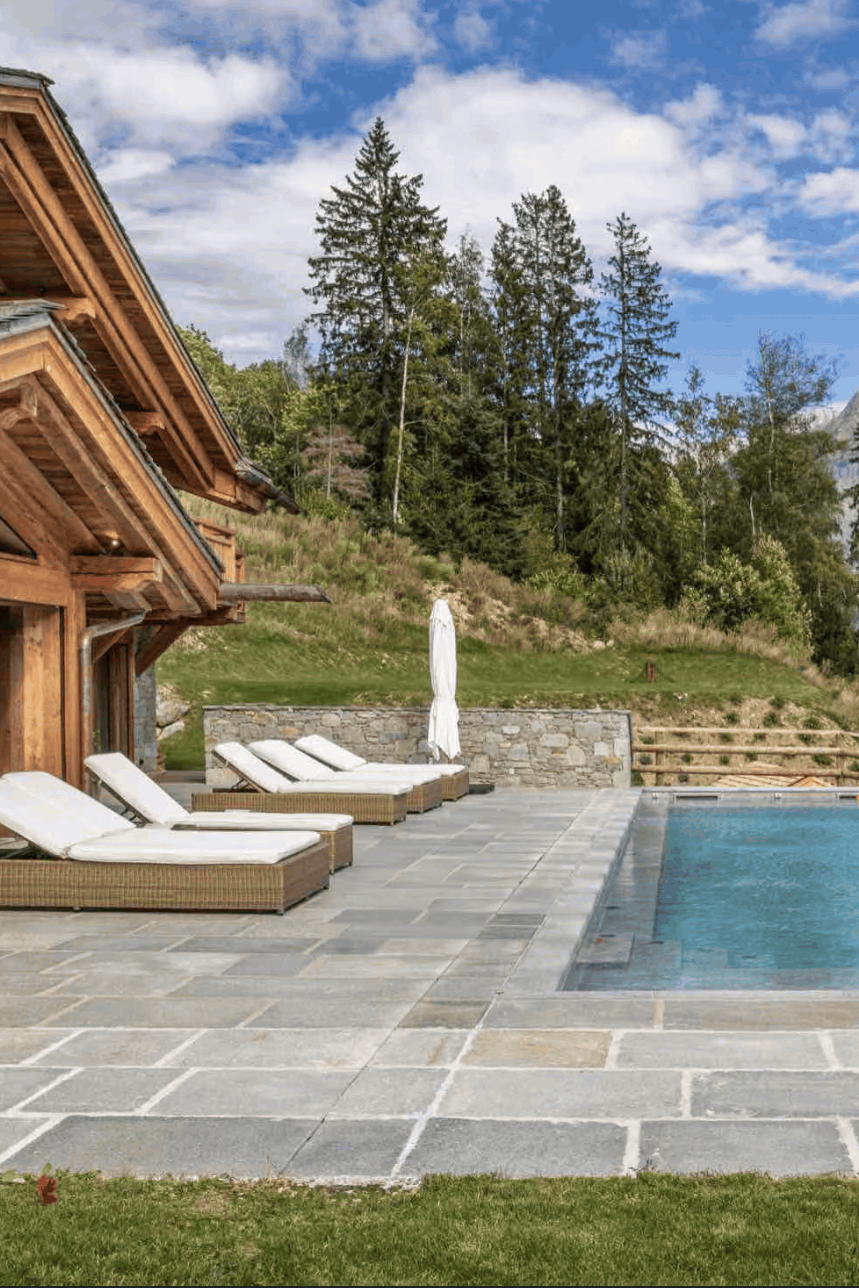 A luxury mountain chalet with a stone patio, outdoor swimming pool, two white lounge chairs, and a stunning backdrop of pine trees and snow-capped peaks under a blue sky.