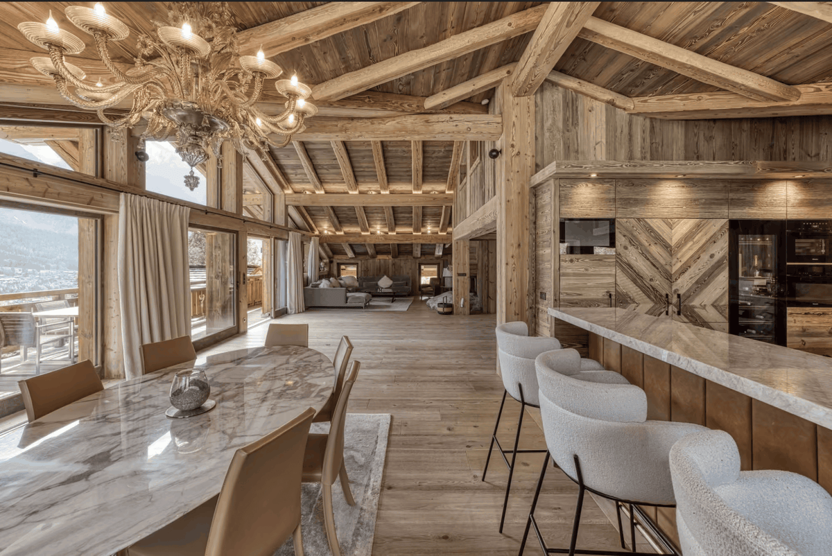A luxury chalet interior featuring a large marble dining table with beige chairs, a high wood-beam ceiling, a marble kitchen island with white bar stools, and a mountain view through the windows.