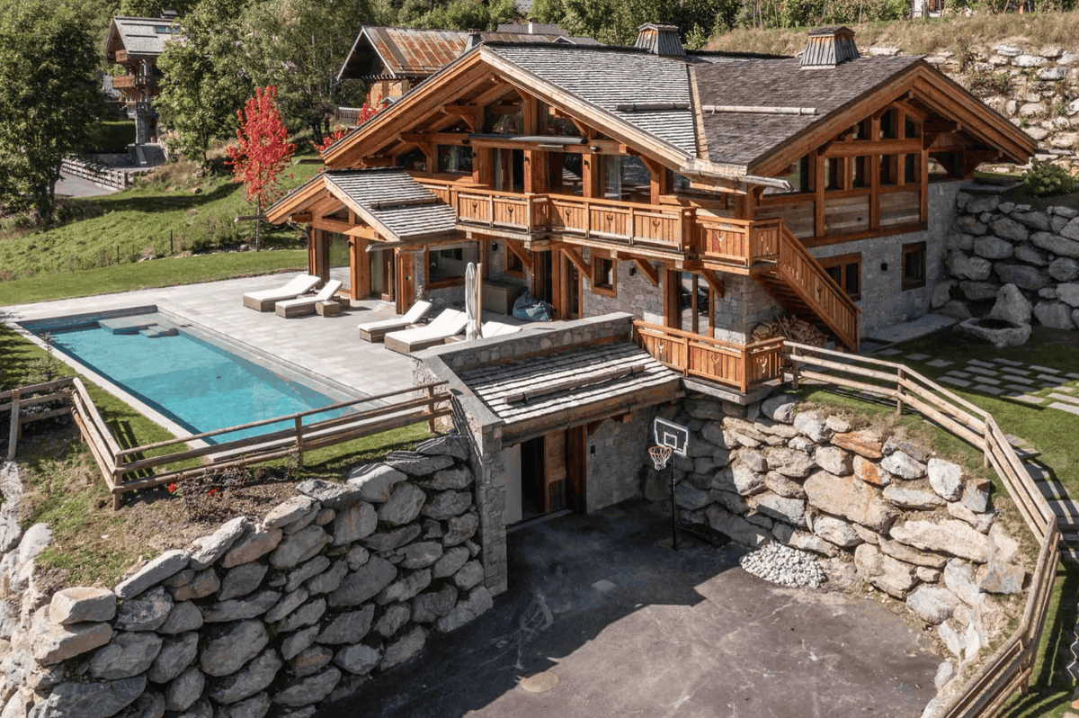 Aerial view of a large alpine-style chalet with wooden balconies, stone walls, a swimming pool, and a driveway, surrounded by trees and landscaping.