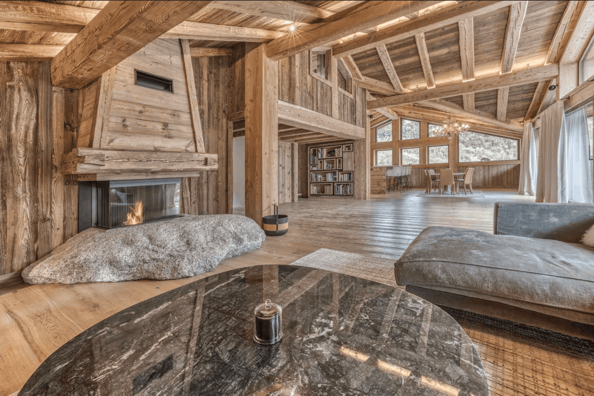 A luxurious rustic chalet living room with a large stone fireplace, a gray fabric chaise lounge, a round dark marble coffee table in the foreground, and an open loft space with wooden beams and windows in the background.