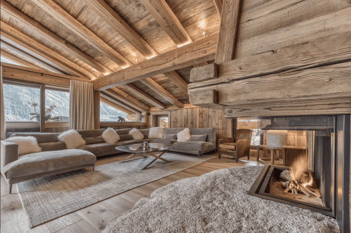 A luxurious mountain chalet living room featuring a large sectional sofa with white sheepskin pillows, a round glass coffee table, a roaring fireplace with a black surround, and a rustic wooden ceiling with exposed beams.