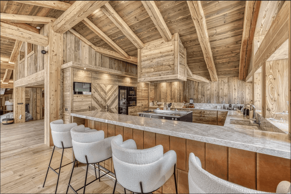 A luxury modern kitchen island in a rustic wooden chalet, featuring a marble countertop, white bar stools, a large wood range hood, and a high vaulted ceiling with exposed wooden beams.