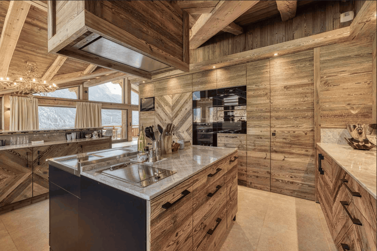 A luxury modern kitchen with rustic wood cabinetry, a central island with a marble countertop and sink, and a large range hood under a wooden ceiling.