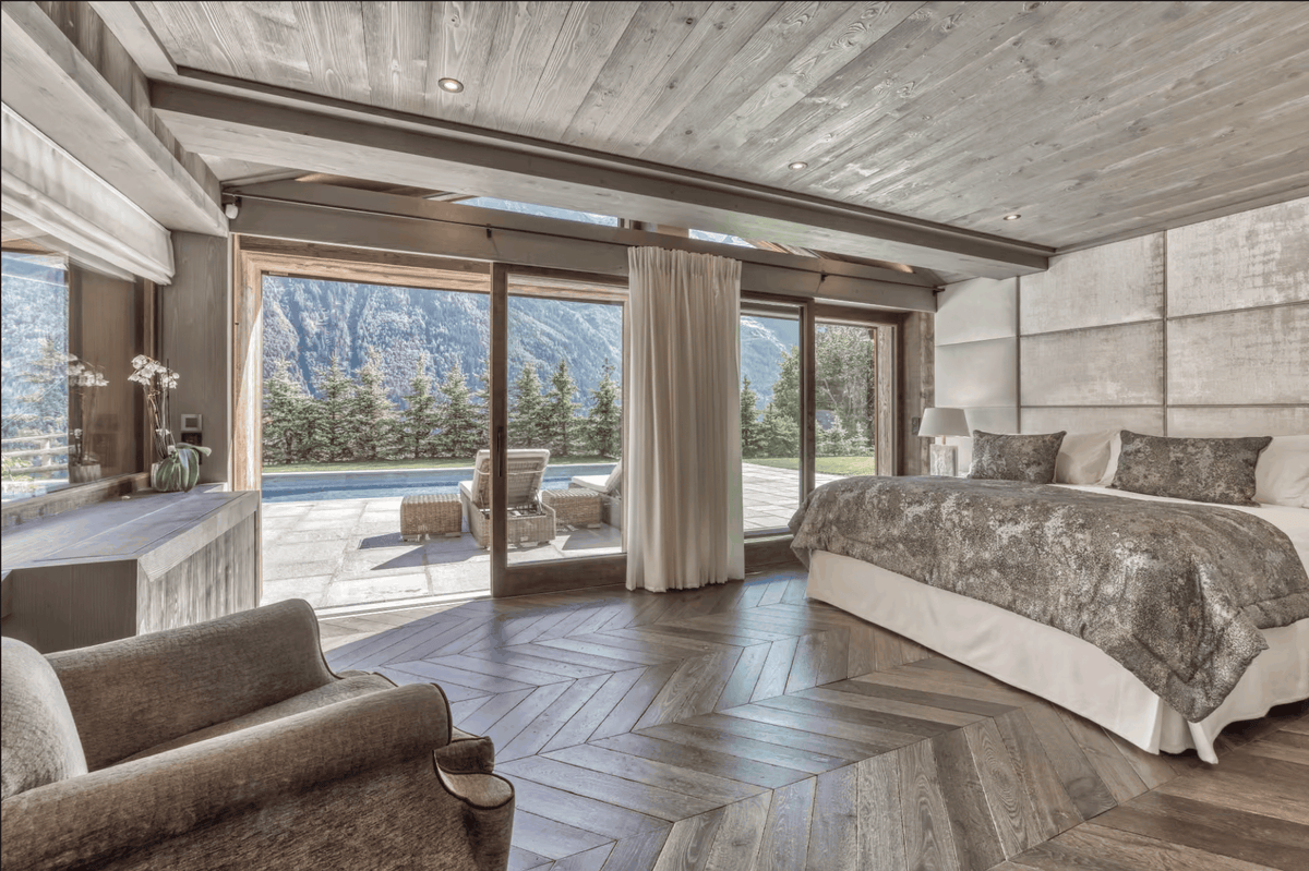 A luxury bedroom interior with a large bed dressed in gray and white patterned bedding, herringbone wood flooring, paneled walls, and floor-to-ceiling windows overlooking a mountain landscape and outdoor patio.