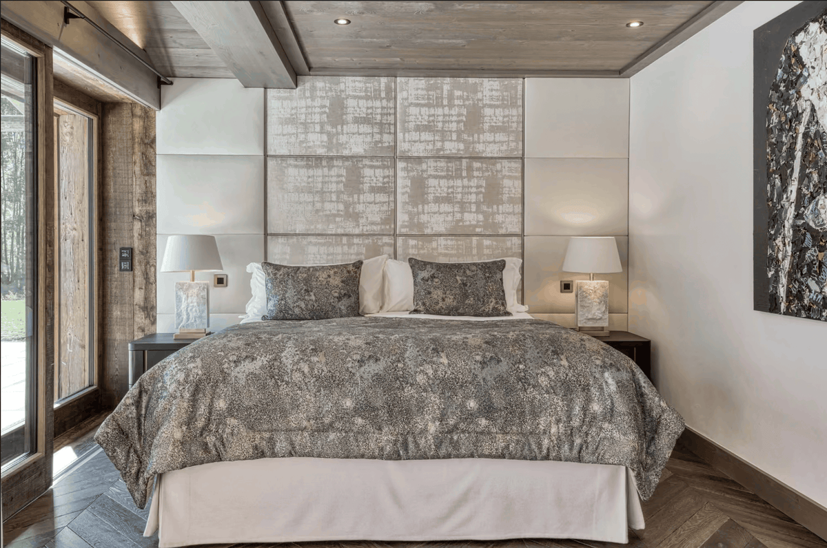 A luxury bedroom featuring a large bed dressed in a textured gray and white patterned comforter and matching pillows, set against a modern white paneled headboard. The room has a rustic wood ceiling, wooden floors, and bedside tables with lit lamps.