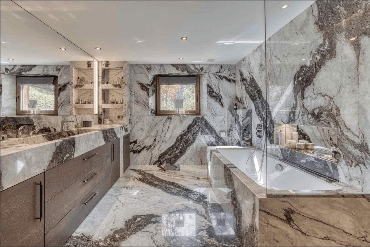 Luxury modern bathroom featuring a double vanity with dark wood cabinets, a white bathtub, and a glass shower screen, all set against dramatic gray and white veined marble walls and floor.
