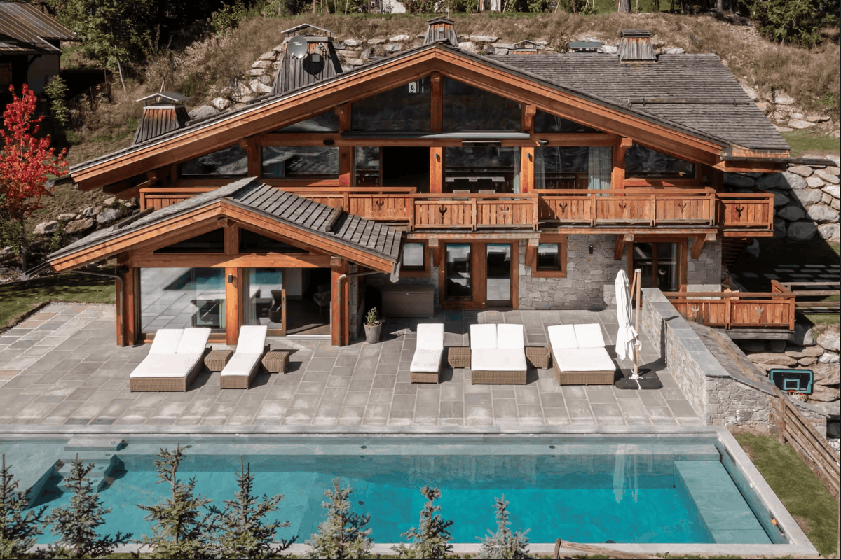 Aerial view of a luxury chalet-style mountain villa with a swimming pool, outdoor lounge chairs, wooden balconies, and a stone and wood exterior.