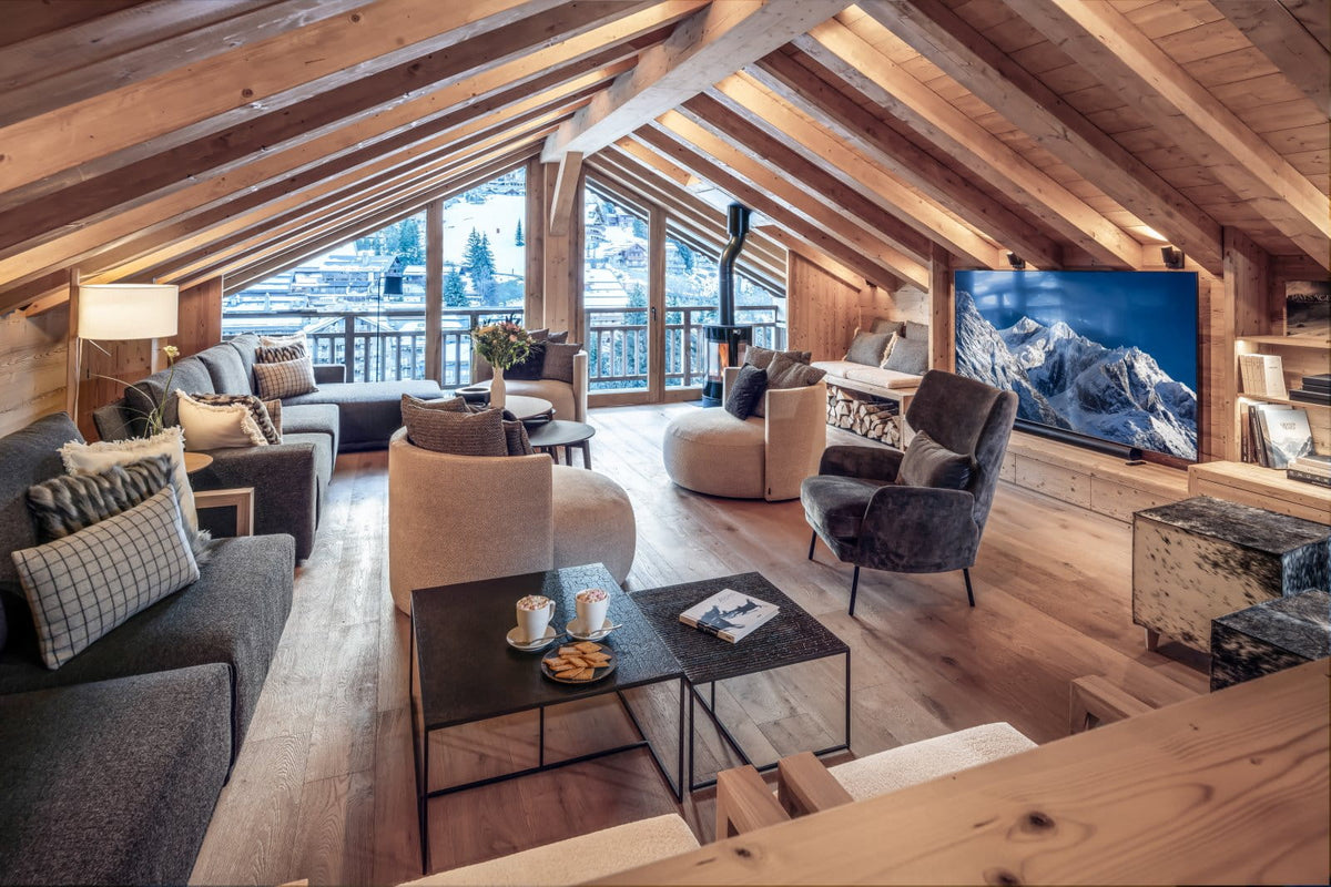 A luxurious ski chalet living room with a large wall-mounted screen displaying a mountain landscape, cozy gray and beige sofas, wooden ceiling beams, and floor-to-ceiling windows.