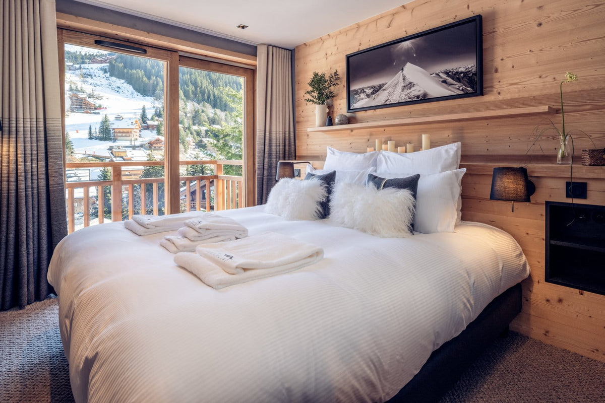 A cozy ski-inclusive bedroom with a large bed dressed in white linens and fluffy pillows, set against a light wood-paneled wall with a framed mountain print. Large windows offer a view of snowy mountains and pine trees.