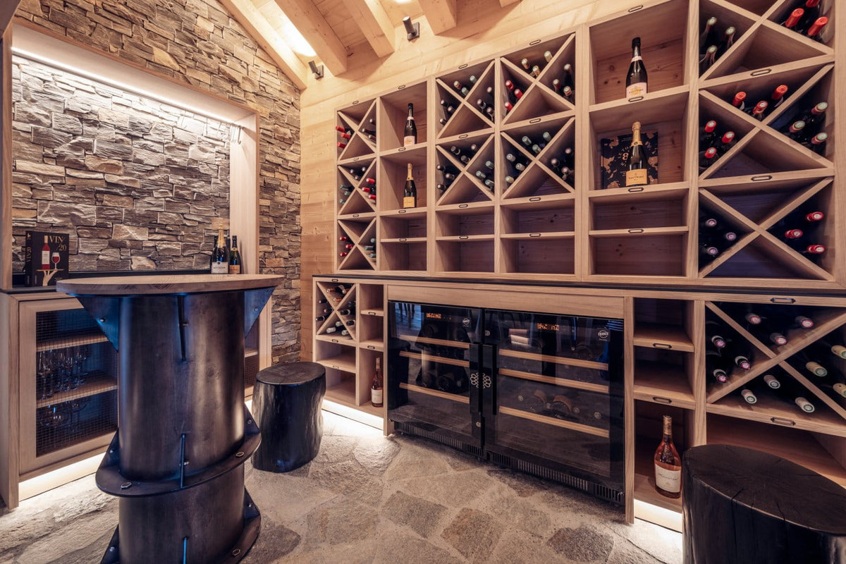 A luxury wine cellar interior featuring a large wooden wine rack system filled with bottles, a stone-tile accent wall, a dark metal bar counter, and two wine coolers under the counter.
