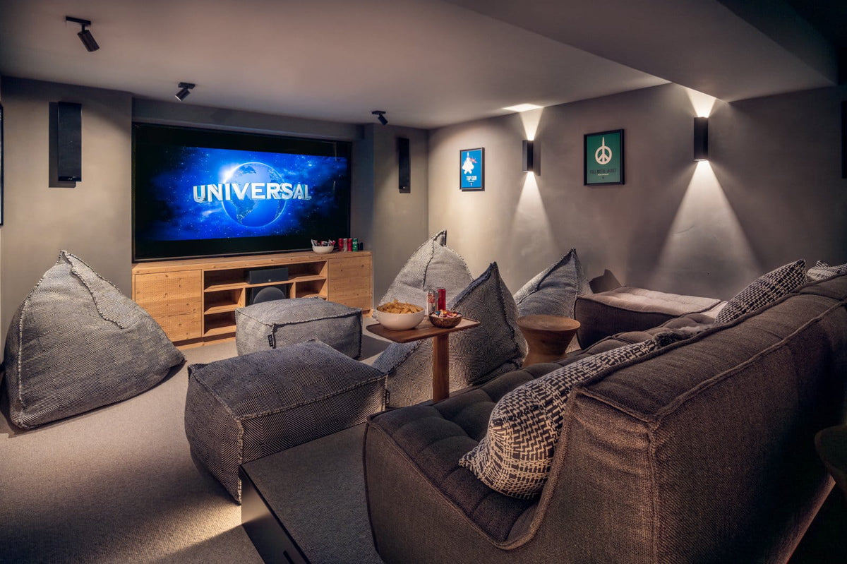 A home theater or lounge room with tiered seating, beanbag chairs, and a large screen displaying the UNIVERSAL logo. The room has gray walls, wall-mounted spotlights, and a wooden media console.