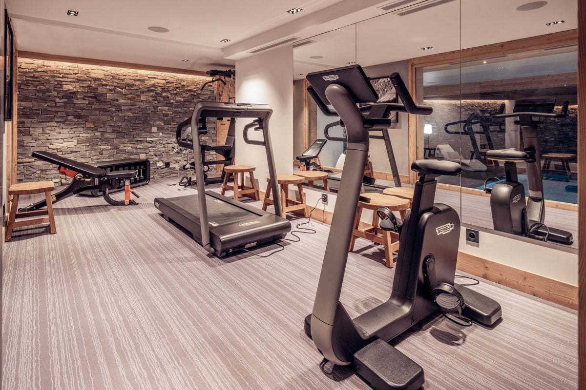 A modern indoor fitness gym with treadmills, an elliptical machine, and a weight bench, set in a spacious room with wood flooring, a stone accent wall, and large mirrors.