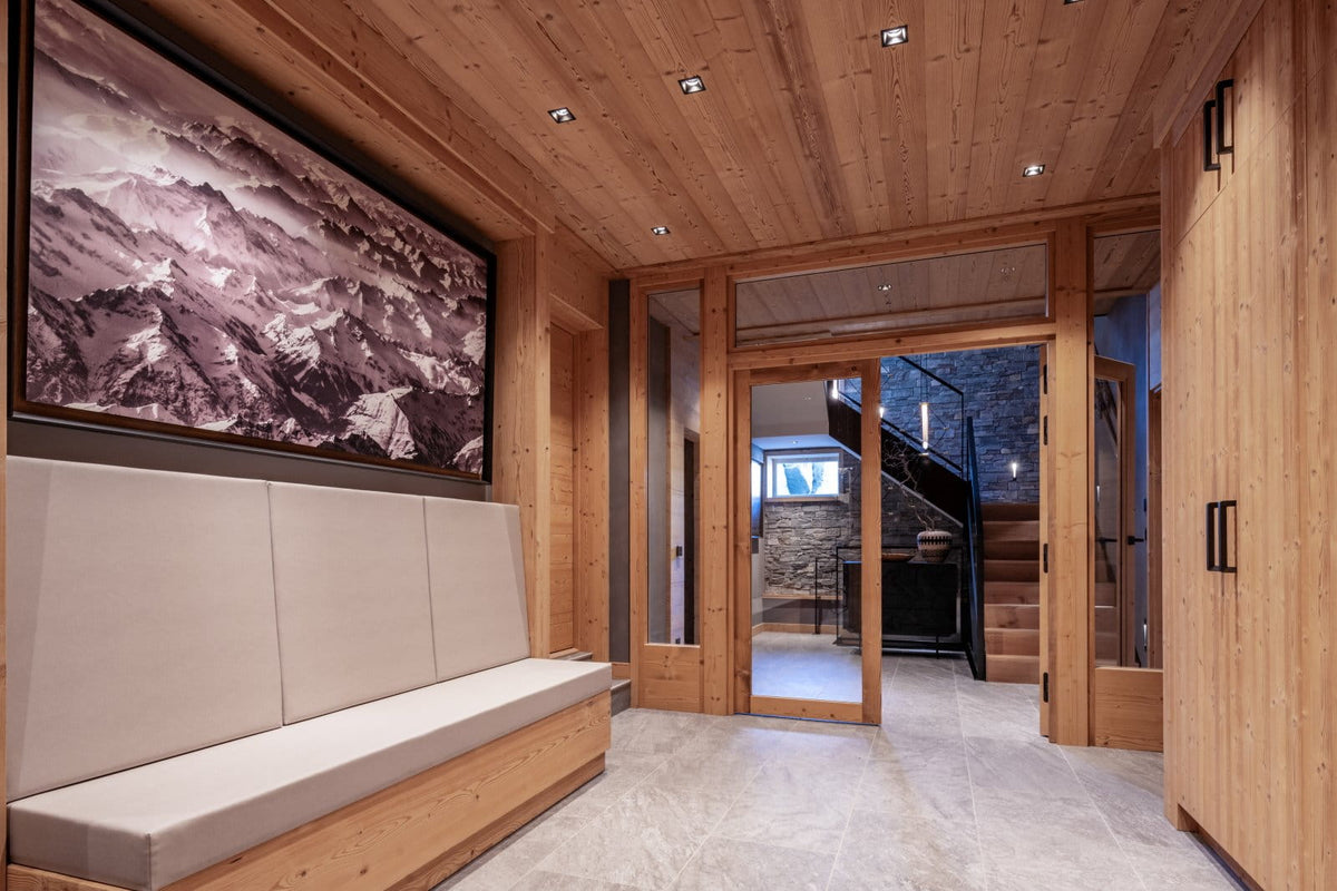 A modern interior hallway with light wood walls and ceiling, a built-in cushioned bench, a large framed mountain landscape artwork on the left, and glass doors leading to a staircase area.