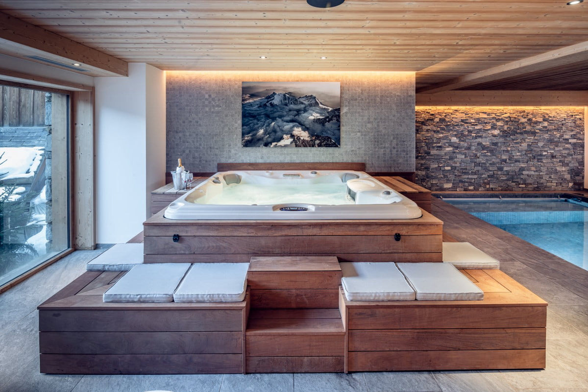 An indoor hot tub and plunge pool area in a modern mountain-style lodge. A rectangular hot tub sits on a raised wooden platform with built-in seating and light cushions, set against a stone and wood wall with a mountain artwork above.