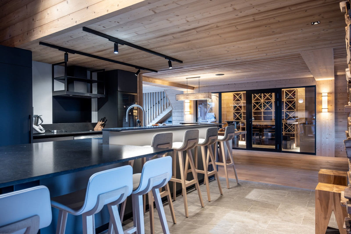 A modern kitchen island with a dark countertop and light wood bar stools, set in a rustic mountain-style home with a wood ceiling and stone floors.