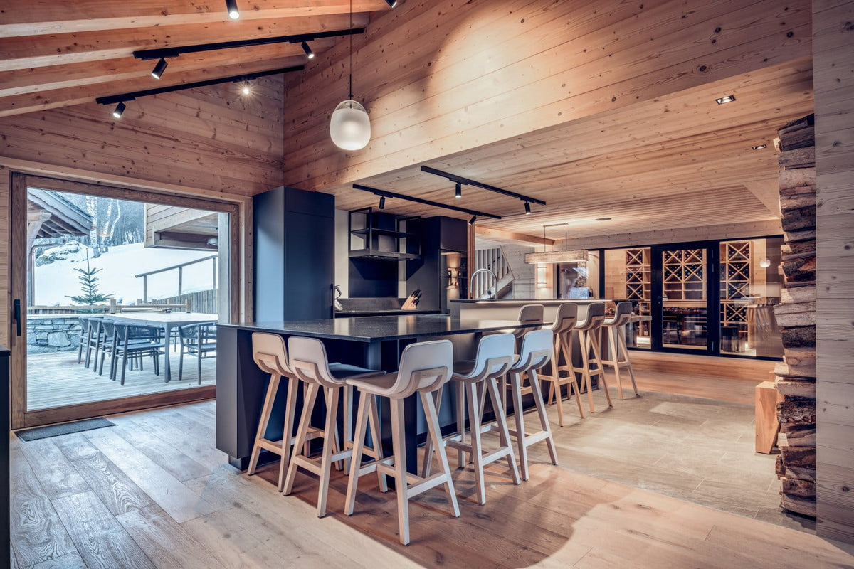 A modern ski chalet interior featuring a long bar with white bar stools, light wood flooring, exposed wood ceiling beams, and large windows looking out onto a snowy mountain landscape.